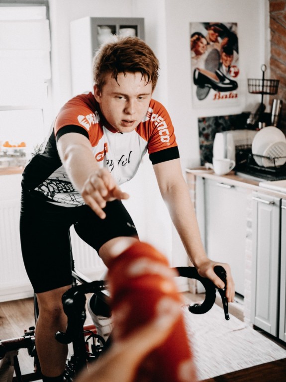 Chlapec v cyklistickém dresu jede vyčerpaný na domácím cyklistickém trenažeru a natahuje se k ruce, která mu podává sportovní lahev Penco.