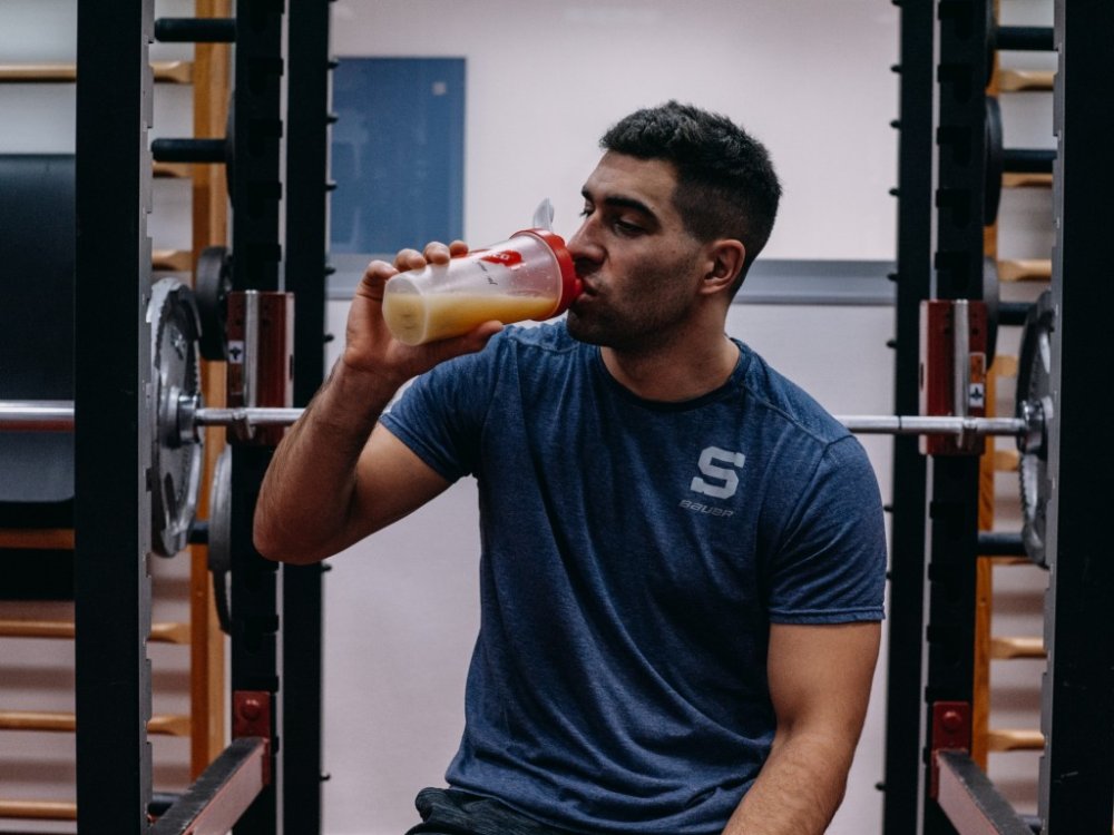 Muž sedí na posilovacím stroji a popíjí protein z šejkru Penco.