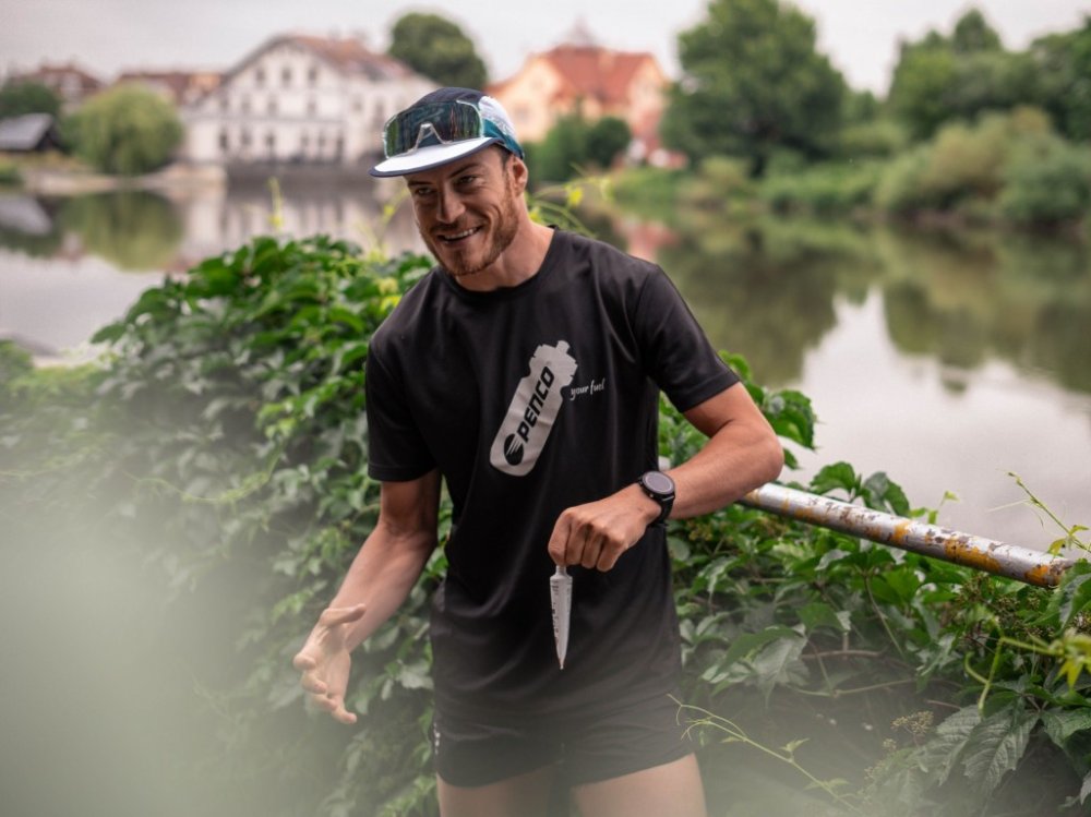 Pavel Wohl v černém tričku Penco stojí u zábradlí nad řekou a v ruce má energetický gel Penco LONG TRAIL v tubě.