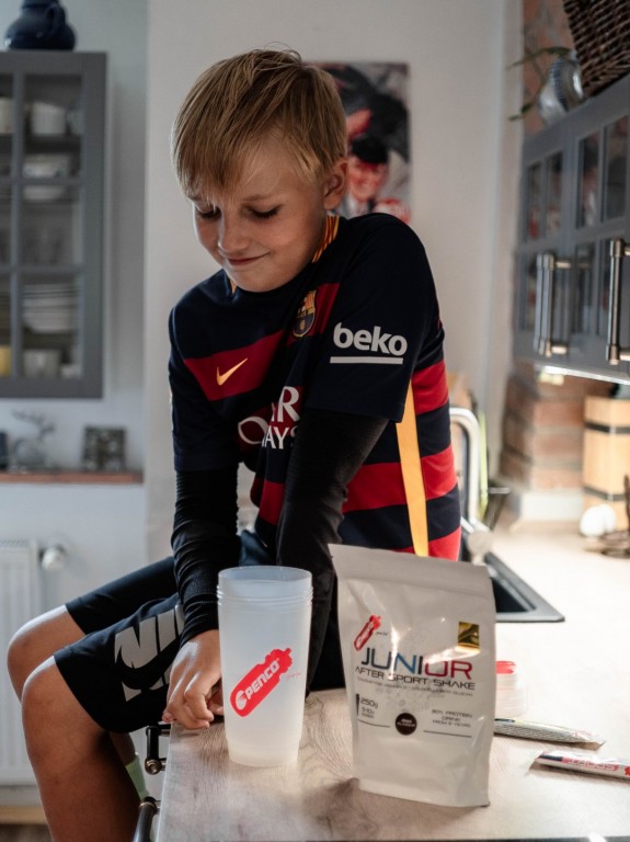 Mladý chlapec v dresu sedí na kuchyňské lince a dívá se na šejkr Penco. Vedle něj na lince stojí bílé balení nápoje Penco Junior After Sport Shake.