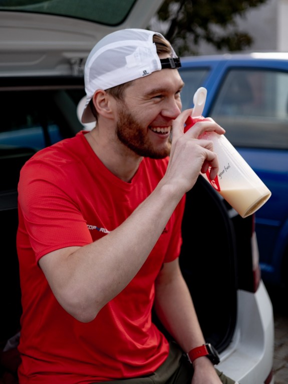 Sportovec v červeném triku a bílé kšiltovce s kšiltem dozadu sedí v kufru auta a popíjí protein z šejkru Penco.