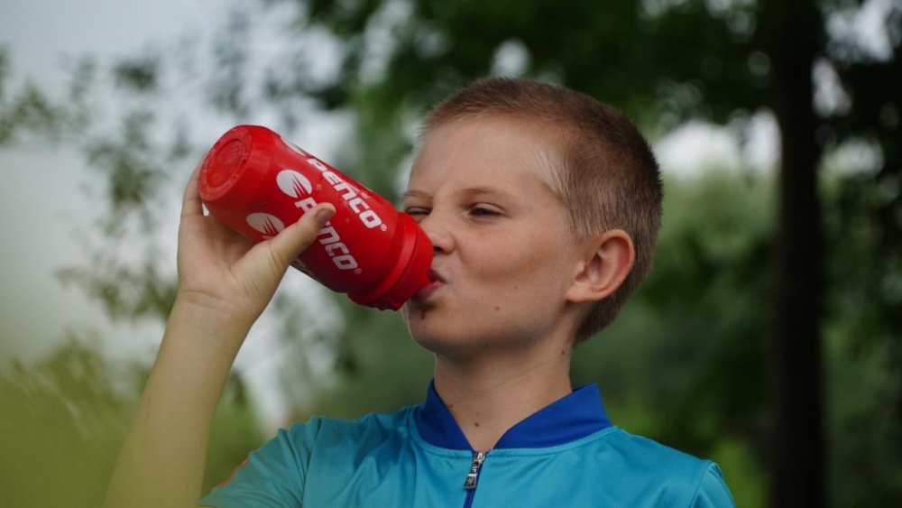 Detailní záběr na chlapce v tyrkysovém sportovním dresu, který venku pije z červené lahve Penco. V rozostřeném pozadí jsou vidět zelené stromy.