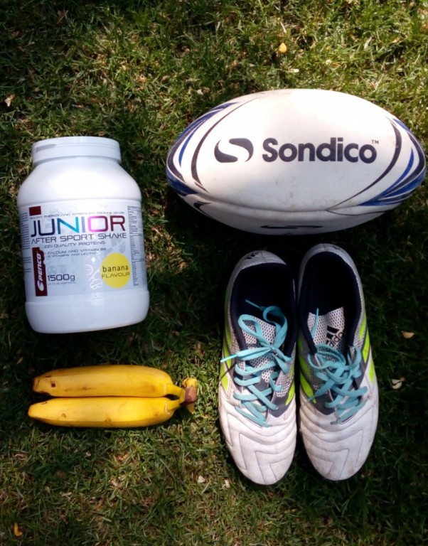 Předměty vyskládané na zemi na trávě: produkt Penco Junior After Sport Shake, 2 banány, bílé sportovní boty a bílý oválný míč.