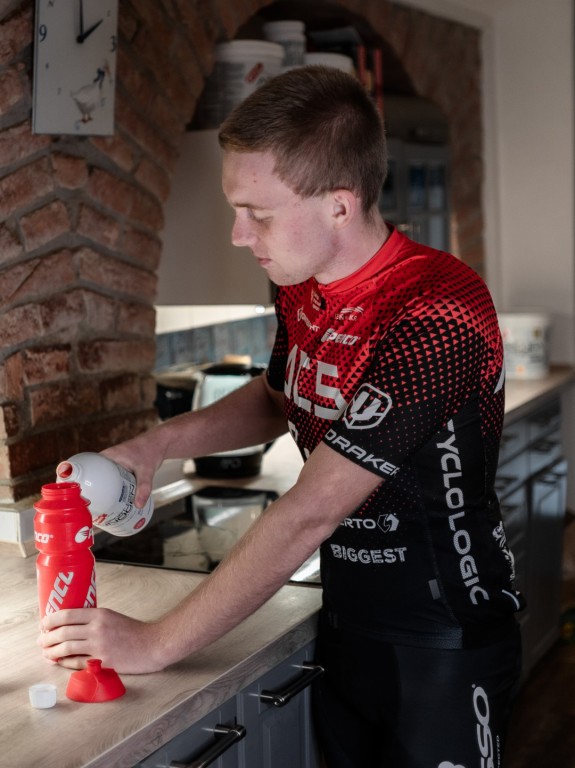 Mladý muž v cyklistickém dresu si doma připravuje sportovní výživu Penco do červené lahve od stejné značky.