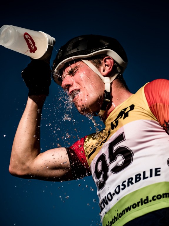 Záběr z podhledu na cyklistu se startovním číslem 95, který si v plném nasazení vylévá vodu z průhledné lahve Penco přímo na obličej. Kolem hlavy mu stříkají kapky vody.