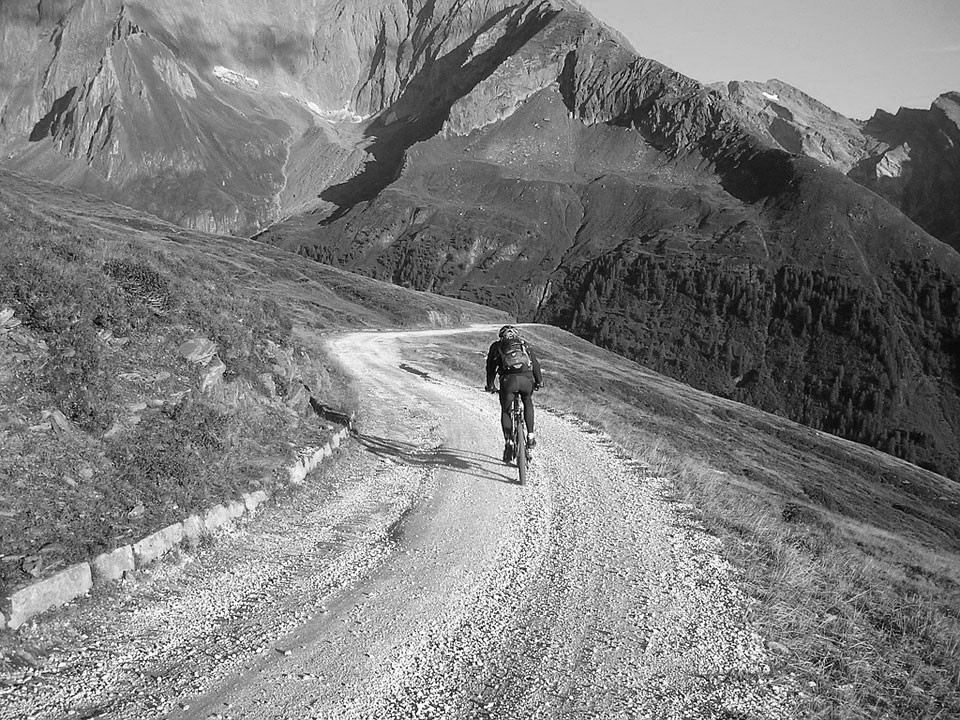 Černobílá fotografie cyklisty, který projíždí silničkou v horách.