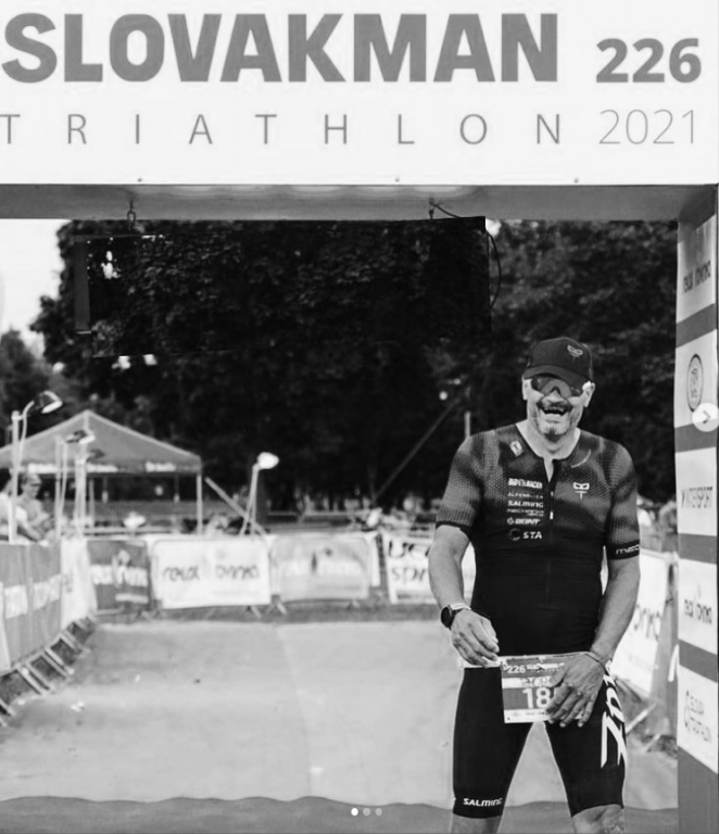 Černobílá fotografie Vladimíra Hrabce, který stojí usměvavý v cílové bráně závodu Slovakman Triathlon 2021. Má na sobě triatlonový dres, kšiltovku a na opasku viditelné startovní číslo.