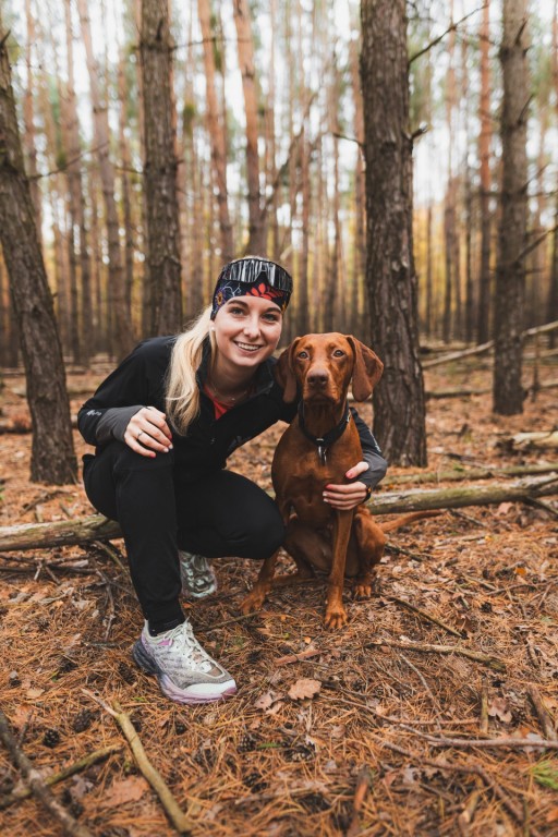 Anička Cylková oblečená ve sportovním oblečení sedí v podřepu v lese a levou rukou objímá svou fenku Ruby.