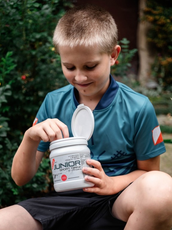Mladý sportovec v tyrkysovém cyklistickém tričku a černých kraťasech sedí venku a s úsměvem otevírá bílou dózu Penco Junior Sport Drink.