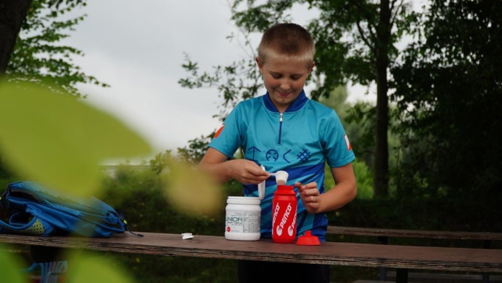 Mladý sportovec v tyrkysovém cyklistickém dresu stojí venku u dřevěného stolu a pomocí odměrky sype prášek do červené láhve Penco. Na stole vedle něj stojí otevřená dóza Penco Junior Sport Drink.