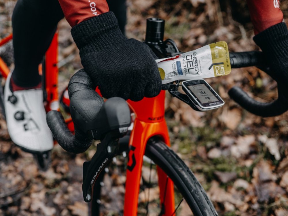 Detailní záběr na ruce v černých rukavicích na řidítkách kola, přičemž pravá ruka zároveň drží energetický gel Penco.