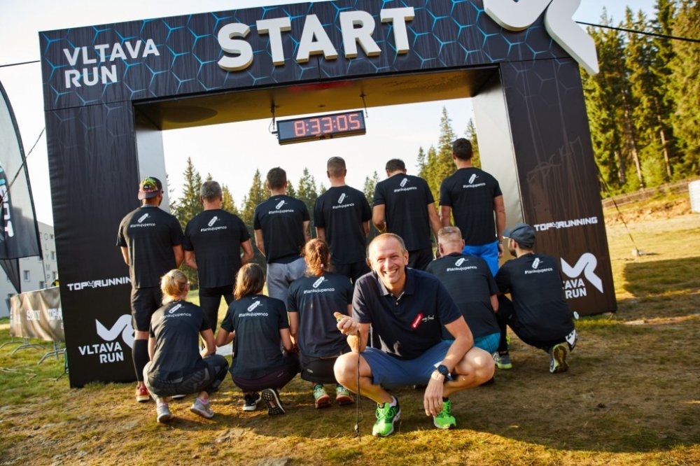 Skupinová fotografie týmu Penco pod velkou černou nafukovací bránou s bílým nápisem 'VLTAVA RUN START'. Většina běžců stojí zády k objektivu a ukazuje černá trička s bílým nápisem '#tankujupenco' a grafikou sportovní lahve. V popředí sedí v dřepu usmívající se muž v modrém polo tričku a v ruce drží stopky. Nad bránou svítí digitální časomíra s červeným údajem 8:33:05.