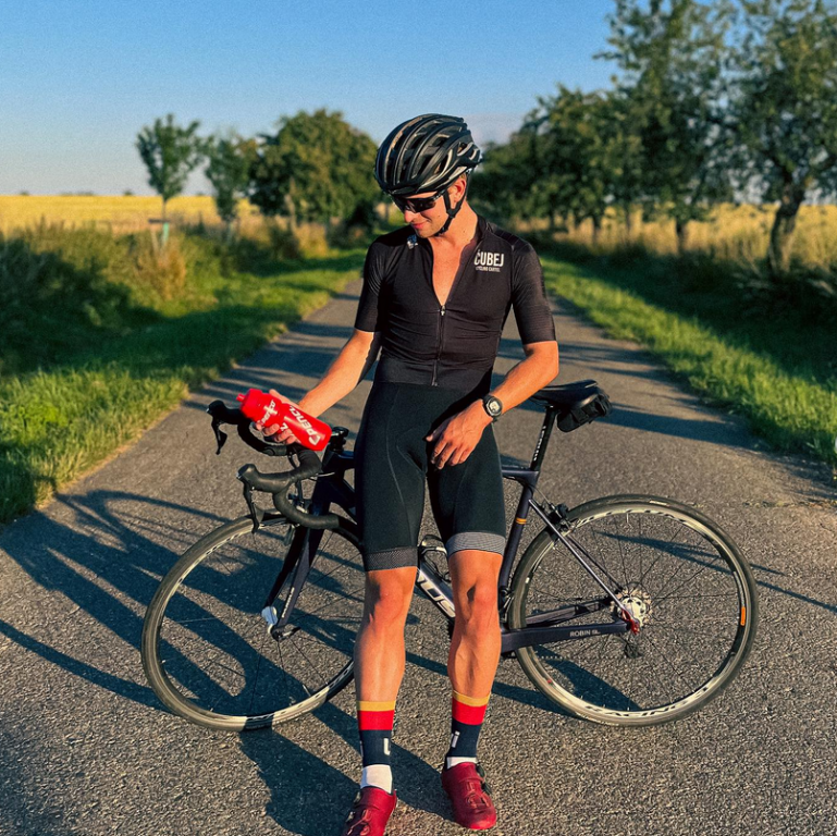 Mladý cyklista stojí na venkovské silnici vedle silničního kola, v ruce drží červenou láhev na pití a dívá se dolů. 