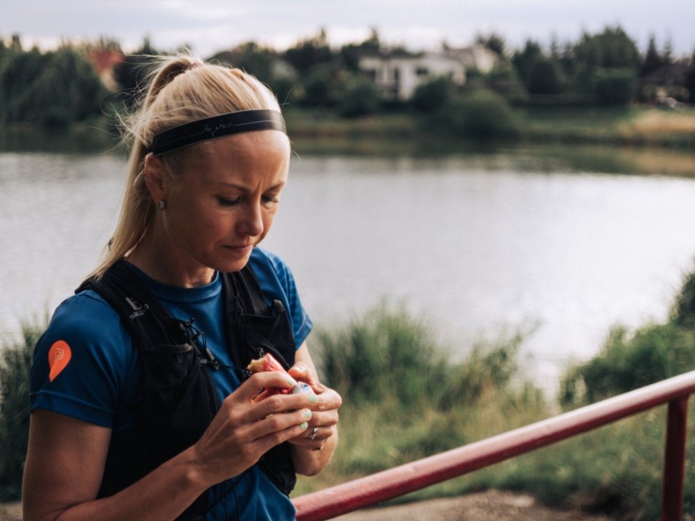 Běžkyně s blond vlasy v culíku stojí u břehu rybníka a rozbaluje energetickou tyčinku Penco. Má na sobě modré sportovní tričko a černou běžeckou vestu.