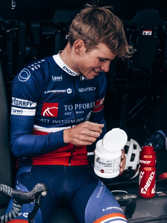 Mladý cyklista v modro-červeném dresu sedí v autě a s úsměvem otevírá bílou dózu s regeneračním nápojem RESTART DRINK. Vedle něj stojí červená cyklistická láhev Penco.
