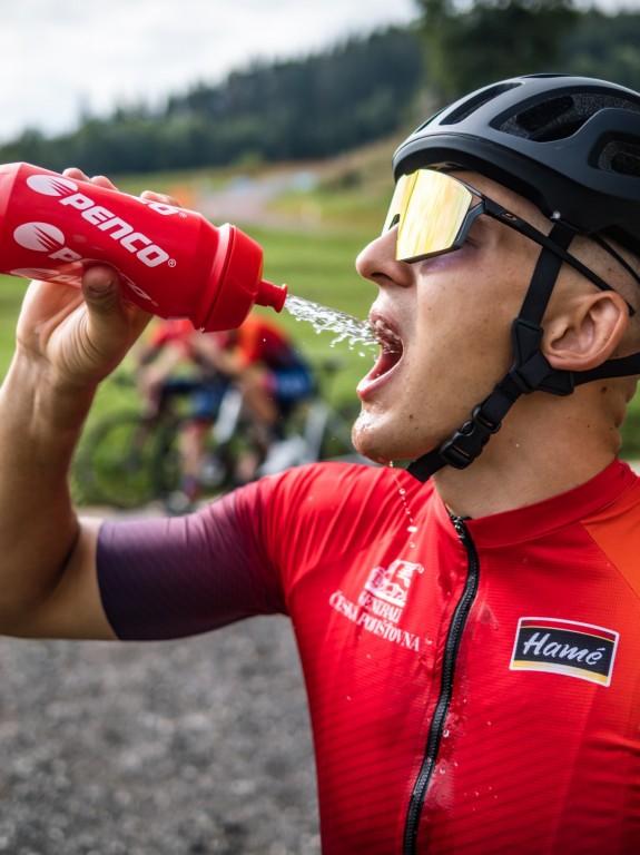 Detailní pohled z profilu na cyklistu v červeném dresu, který si z červené lahve Penco stříká vodu přímo do úst. Má na sobě černou helmu a sportovní brýle se zlatým zrcadlovým zorníkem.