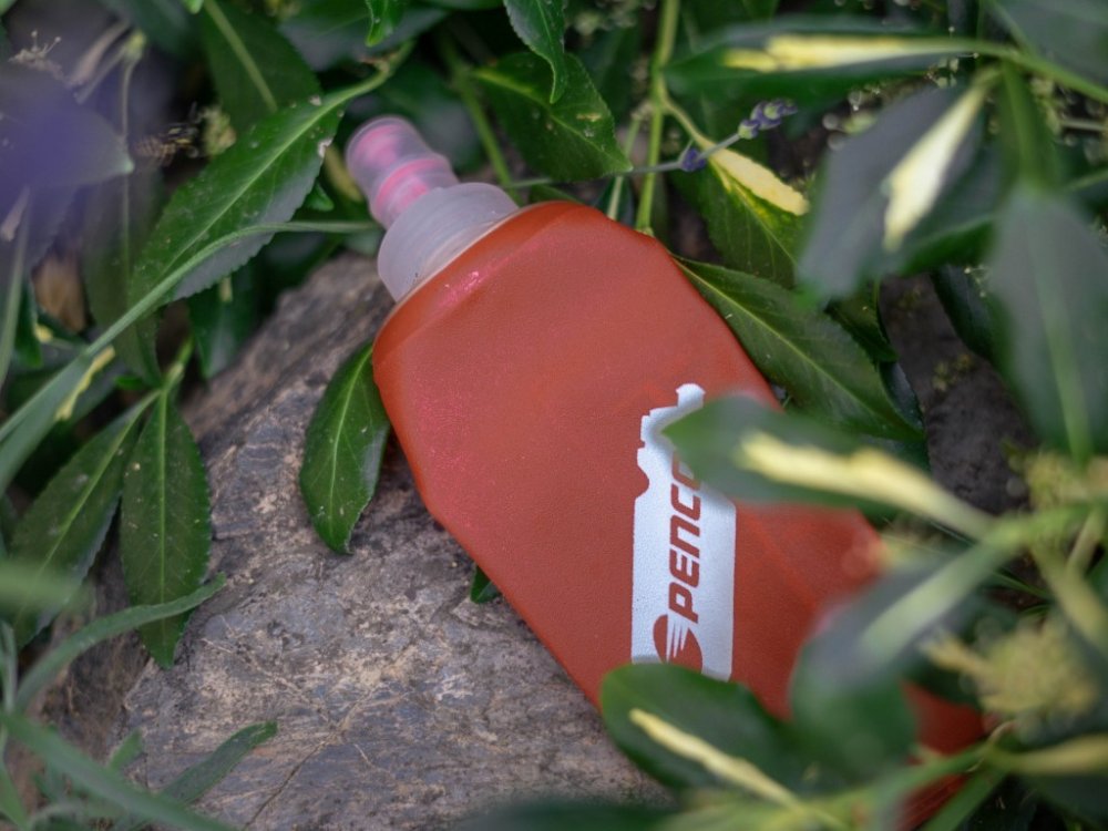 Soutěžní malá červená Penco lahvička (soft flask) položená na kameni a částečně zakrytá zelenými listy.