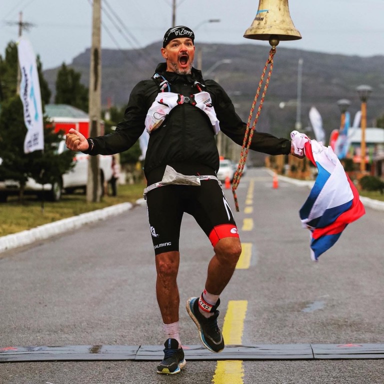 Triatlonista doběhl do cíle, v levé ruce drží složenou vlajku a křičí radostí.