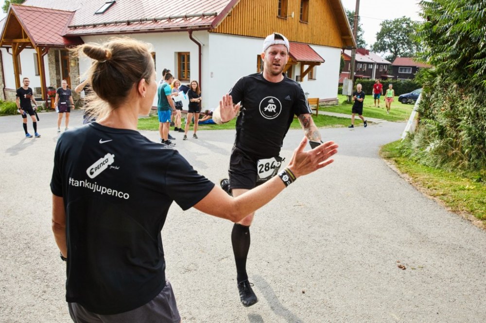 Pohled zezadu na běžkyni v černém tričku s nápisem '#tankujupenco' a grafikou sportovní lahve, která má připravenou nataženou ruku na placáka (high-five) s běžcem v černém tričku se startovním číslem 284, který proti ní běží na asfaltové silnici.