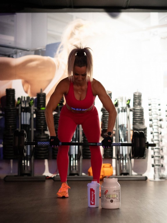 Žena posiluje v posilovně a má před sebou na zemi produkty sportovní výživy