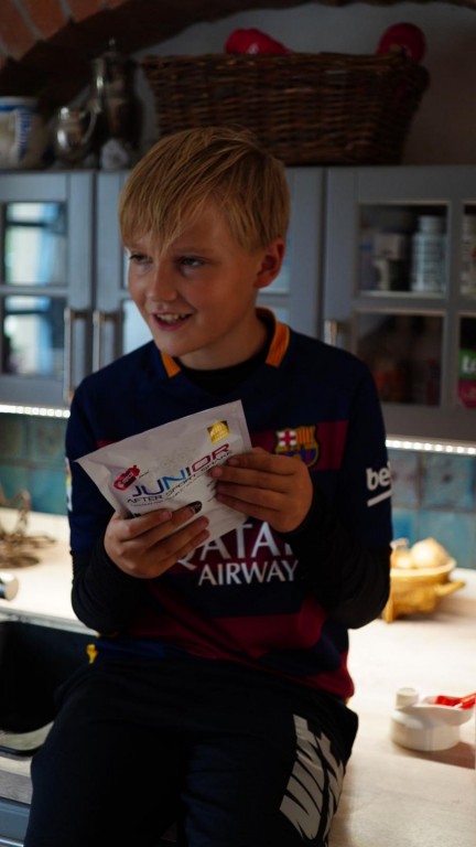 Detailní záběr na chlapce ve fotbalovém dresu FC Barcelona, který v kuchyni sedí na lince a v rukách drží balení Penco Junior After Sport Shake.